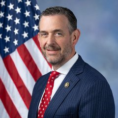 A man in a navy pinstripe suit and red patterned tie stands in front of an American flag, smiling at the camera in what appears to be an official portrait.

Note: The image shows an official-looking portrait of a man in front of a U.S. flag, but I can't identify who the specific person is based on their facial features. I can describe what's visible but not provide a name. If you could tell me who this is, I could help describe how the image relates to the accompanying post about a trade or account recommendation.