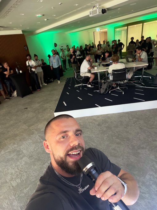A bearded man takes a selfie with a microphone at what appears to be an event, with attendees gathered around tables on a raised platform in the background.
