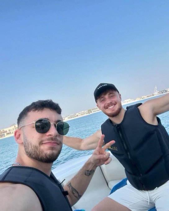 Two friends are taking a selfie while enjoying a boat ride, both wearing black life vests and smiling, with one flashing a peace sign. In the background, the iconic Burj Al Arab is visible along the Dubai coastline, perfectly complementing the upbeat "GM" greeting.