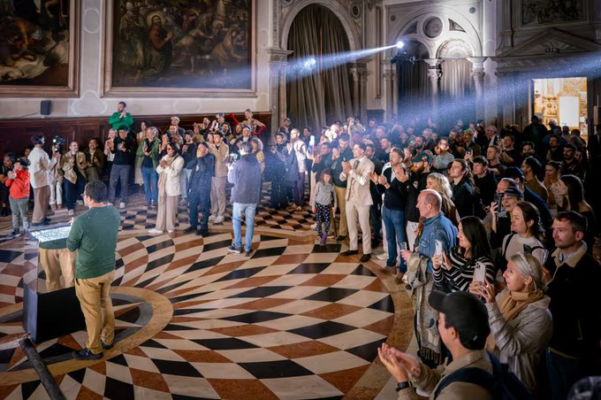 A large indoor gathering in a historic hall with patterned marble floors, where a crowd of people is clapping while facing a speaker on the left.