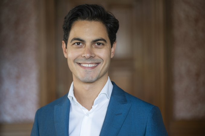The image features a smiling man wearing a blue suit jacket and white shirt who appears happy and focused, likely posing for a photo or engaging with the surrounding content related to Bitcoin. However, there are some technical details visible in the background that might be relevant to discussions about cryptocurrency tax laws. It is unclear whether these details have any connection to the positive tone of the image, as they may not directly relate to the man's expression and pose. The overall atmosphere seems to be one of discussion or awareness related to the effects of certain government regulations on Bitcoin.
