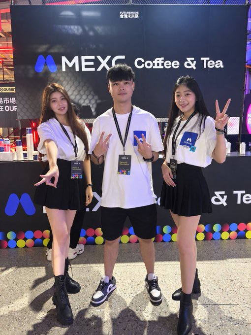 Three young people pose in front of a "MEXC Coffee & Tea" booth at what appears to be a crypto/blockchain event, with one man in the center wearing a MEXC lanyard between two women making hand gestures.