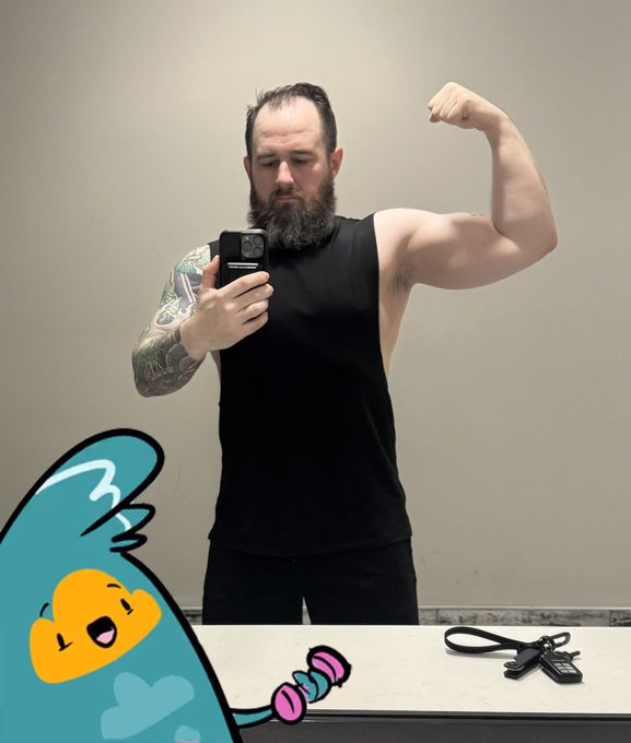 A bearded man in a black sleeveless shirt takes a mirror selfie while flexing his arm, with a cartoon character holding dumbbells edited into the bottom left corner of the image.