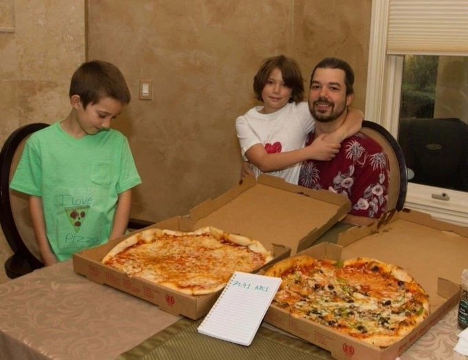 A man and two boys smiling behind two open pizza boxes on a table, with a notepad displaying 'PEKI AKA' placed in front.