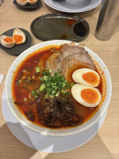 A bowl of spicy ramen topped with sliced pork, a halved soft-boiled egg, green onions, and black wood ear mushrooms in a red broth.