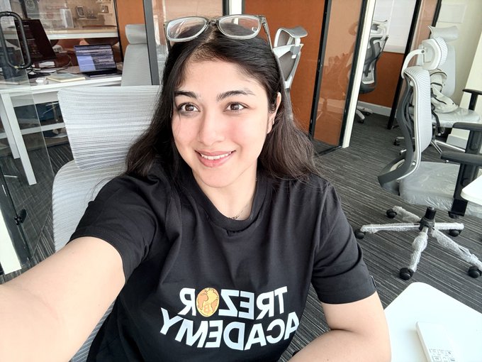 A smiling woman takes a selfie while wearing sunglasses on her head and a black t-shirt displaying the upside-down text "TREZOR ACADEMY" alongside a Bitcoin logo, in a modern office space with ergonomic chairs and laptops in the background. The image reflects her excitement for the upcoming BitcoinHer21 phase 2 event in Bengaluru, where Trezor Academy is partnering to deliver a hands-on Bitcoin learning experience.