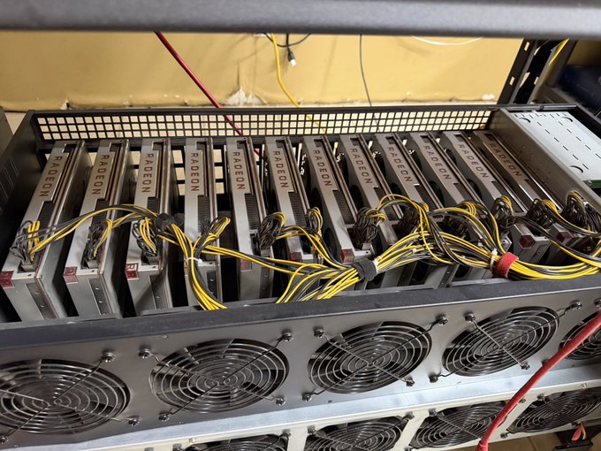 A photograph of a cryptocurrency mining rig containing multiple Radeon graphics cards connected by yellow and black power cables, with cooling fans visible below.