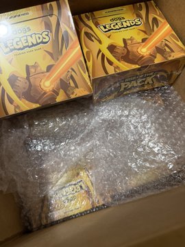 An opened shipping box containing bubble wrap and two yellow 'Yu-Gi-Oh! LEGENDS Booster Pack' boxes with artwork of a warrior character.