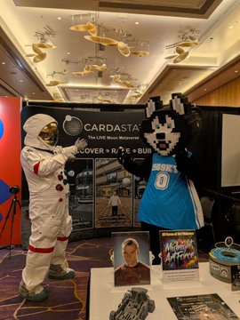 Two cosplayers pose in front of a Cardasta merchandise banner at a convention, one in an astronaut suit and the other in a mascot costume wearing a blue Orlando Magic basketball jersey, with related merchandise displayed on a table.