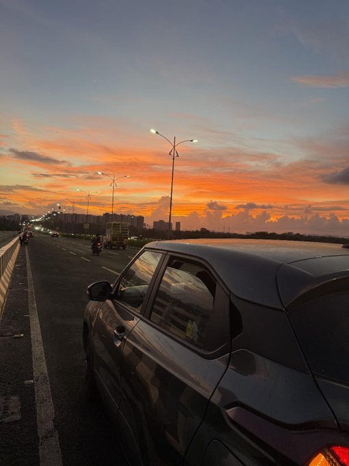 Scenic photograph of a silver car parked on a bridge at sunset, featuring a vibrant orange and purple sky over a body of water with city buildings in the distance.