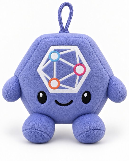 A small purple hexagonal plush toy with a smiling face, carrying a loop at the top and decorated with a white geometric design featuring colored circles.