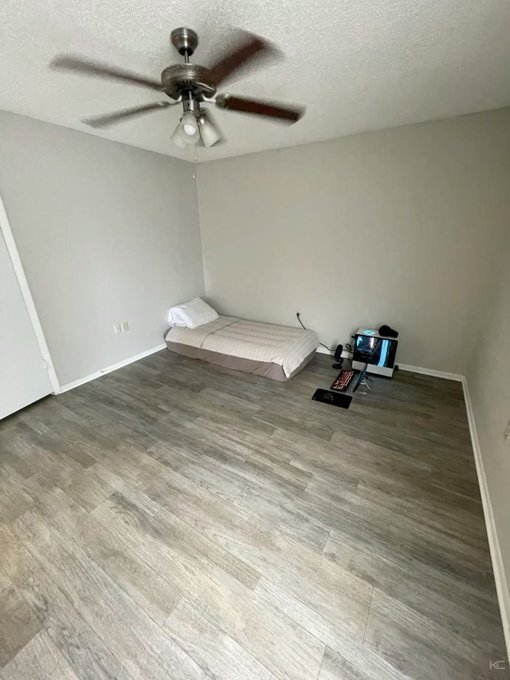 An empty bedroom with gray wood-style flooring, gray painted walls, a ceiling fan with lights, and a simple mattress placed directly on the floor in the corner with a small TV on the floor nearby.