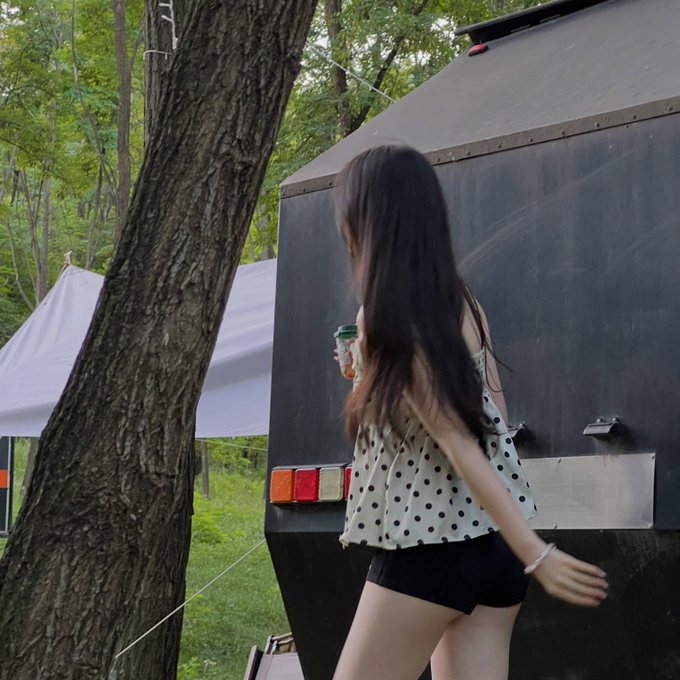 A photograph of a woman with long dark hair, wearing a polka dot top and black shorts, holding a drink can and walking past a black trailer in a wooded outdoor area.