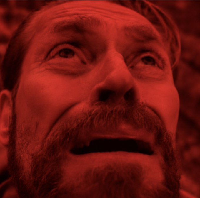 A close-up of a bearded man's face bathed in red illumination, looking upward with his mouth slightly open.