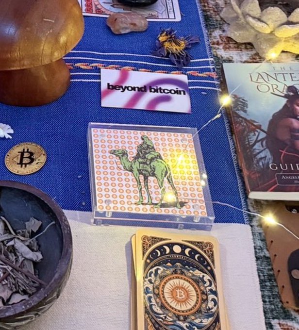 An arrangement featuring a "beyond bitcoin" card, a deck of Bitcoin-themed playing cards, and a card depicting a figure on a camel, set amongst crystals and string lights.