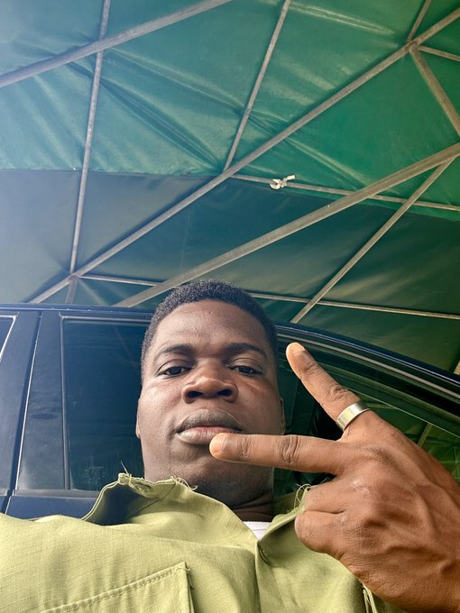 A low-angle selfie of a man in an olive green shirt making a peace sign, wearing a gold ring, photographed under a green canopy roof.