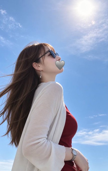 A woman with long brown hair, wearing sunglasses, a red top, and a cream cardigan, poses against a bright blue sky with the sun shining behind her.