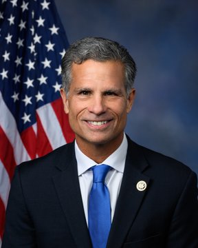 A smiling man in a dark suit with a blue tie poses in front of an American flag, wearing what appears to be a congressional pin on his lapel. The image has the polished look of an official government portrait, fitting a post asking "How we feeling about this?"