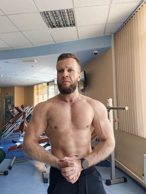 A shirtless, muscular man with a beard stands in a gym setting, posing with his hands clasped in front of him.