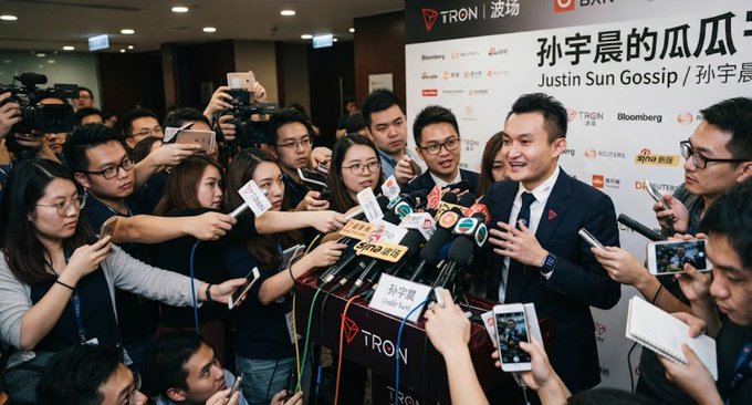 A press conference photo showing Justin Sun (孙宇晨) smiling and being interviewed by a crowd of journalists holding microphones and smartphones, with the TRON/波场 logo and Bloomberg backdrop visible behind him.