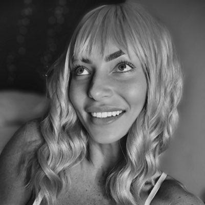 Black and white close-up portrait of a smiling young woman with long wavy blonde hair and bangs.
