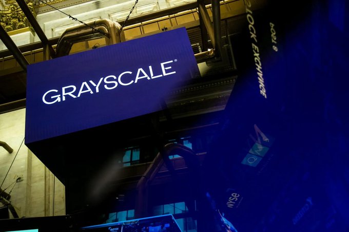 An illuminated "GRAYSCALE" sign hangs from the ceiling of an industrial-looking space, with partial "BLOCK ETFS" text visible on a screen beside it. The branding visually represents the crypto asset manager at the center of the post's news about Grayscale withdrawing its proposed ETF filings for Cardano ($ADA), Polkadot ($DOT), and Hedera ($HBAR).