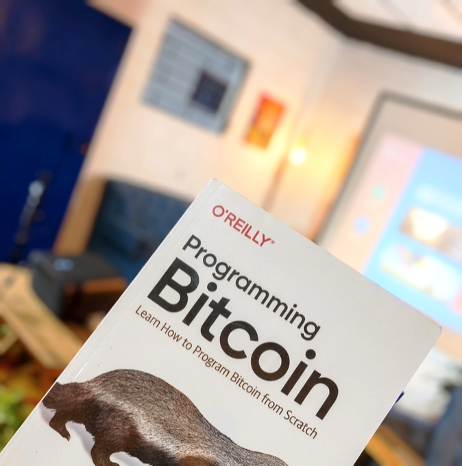 An O'Reilly book titled 'Programming Bitcoin' with the subtitle 'Learn How to Program Bitcoin from Scratch', held up in a room with a TV in the background.
