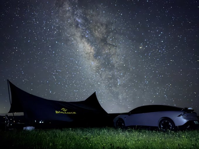 A car parked next to a Binance-branded tent under a stunning Milky Way sky, capturing the Perseid Meteor Shower as part of Binance's Ambassador Program outreach. The "BINANCE" logo is illuminated on the tent, tying the celestial scene to the crypto platform's star-gazing campaign.