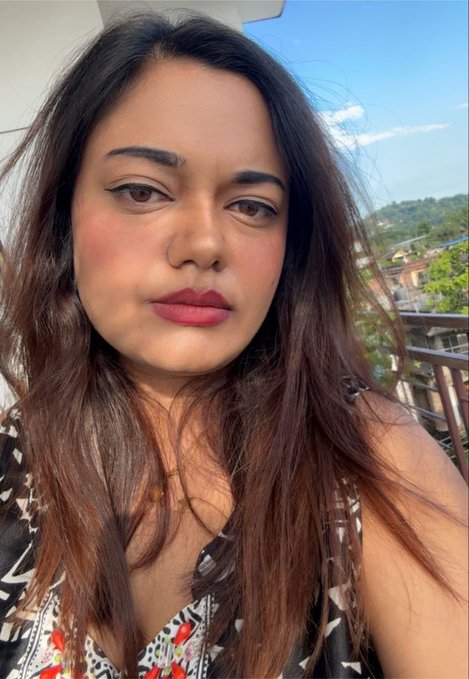A woman with long dark hair takes a selfie outdoors, wearing a patterned top with red accents, with a balcony railing and hillside view in the background.