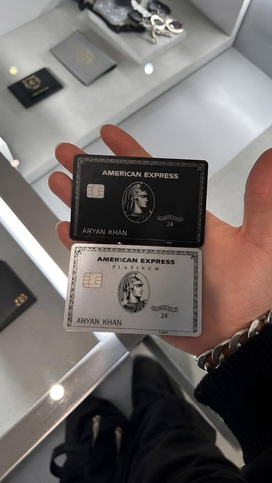 A hand holding two American Express cards (one Centurion 'Black' card and one Platinum card) in the name of ARYAN KHAN, with luxury items visible on a shelf in the background.