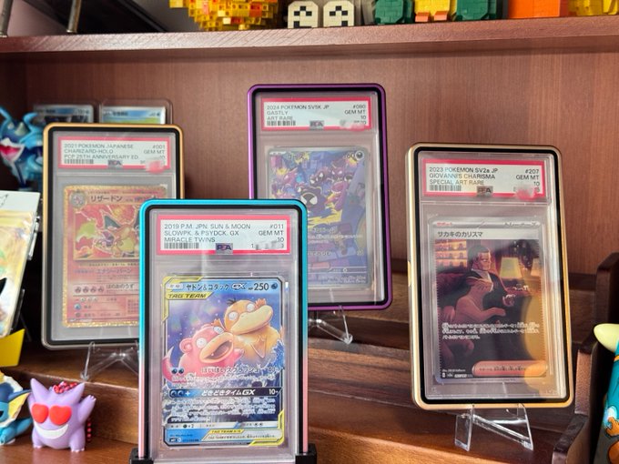 The image shows a collection of four Pokemon cards displayed on a wooden shelf or display case, each card featuring different characters from various games. The cards are arranged in a row with their backs facing upwards, showcasing their vibrant colors and intricate designs. The text "TermMaxFi" is visible in the image, which could be related to the name of the store or the specific Pokemon cards on display.