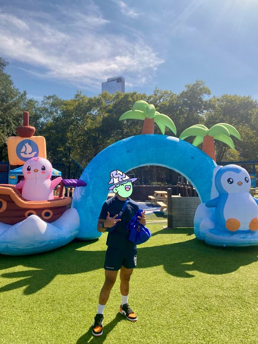 A person in athletic wear and a bucket hat poses in front of a colorful inflatable display featuring cartoon penguin characters, palm trees, and a pirate ship at an outdoor event.