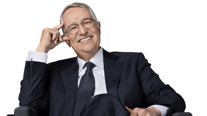 The image features a man wearing glasses and a suit sitting comfortably in an office chair with his legs crossed. He has a smile on his face as he looks directly at the camera, suggesting that he might be having a conversation or posing for a photo. The background of the image is white, which contrasts nicely with the man's dark suit and tie. There are no other objects or people in the image, making it a simple yet striking portrait of the man dressed in business attire.