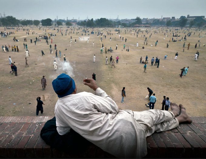 A man wearing a blue turban and white traditional clothing reclines on a brick ledge while watching a large group of people, likely playing cricket, on a vast dusty field.