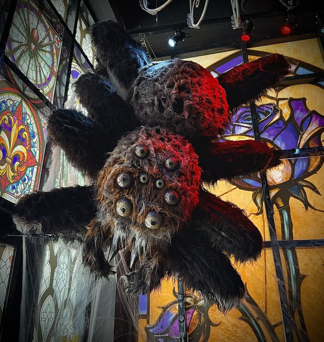 A large, menacing spider-like creature prop with multiple eyes and fangs suspended in front of colorful stained glass windows.