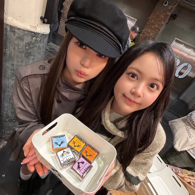 Two young women pose together holding a container of colorful decorated treats, one wearing a stylish newsboy cap, while the caption references "lumen" — perhaps a playful nod to the bright, varied colors of the confections contrasting with moody urban surroundings.