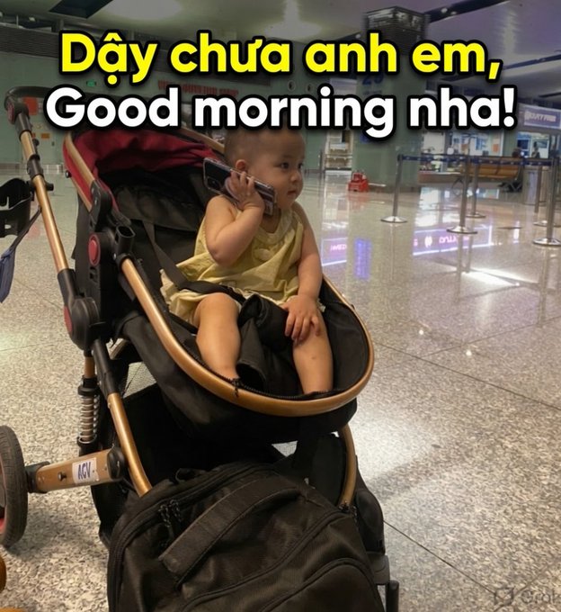 The image features a baby sitting in a stroller, holding a cell phone up to their ear. The background of the photo shows an airport terminal with various signs and equipment visible. The caption of the image reads "Hello anh em! Happy Friday nha anh em! Anh em vào lệnh ơn gì ngon ăn mừng được rồi nhỉ!".