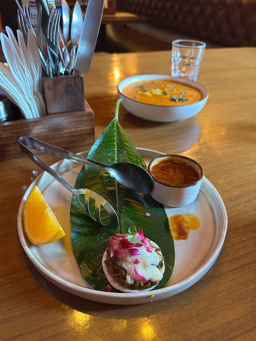 A restaurant dish is elegantly plated on a banana leaf atop a white plate, featuring a small taco-like item with pink garnish, a lemon wedge, and a small bowl of orange sauce, with a bowl of orange soup in the background.