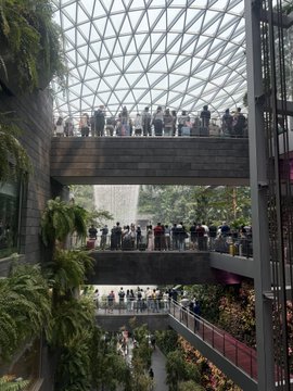 Visitors stand on multiple tiered walkways inside a modern, multi-story building featuring a striking geometric glass ceiling and lush greenery.