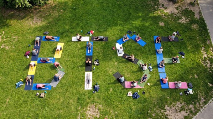 Aerial view of people doing yoga on colorful mats arranged to spell out 'BISO', surrounded by grass.