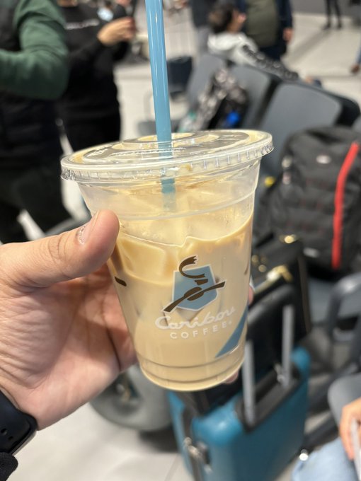 A hand holds a plastic cup of iced coffee with a blue straw from Caribou Coffee in what appears to be an airport waiting area.