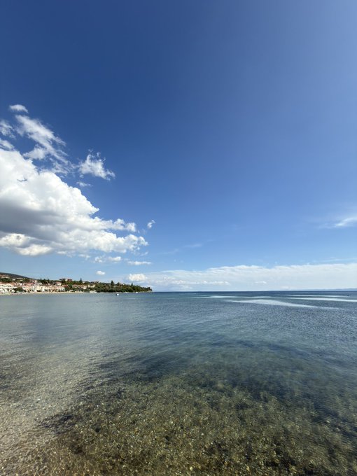 A serene beach scene with crystal-clear turquoise water in the foreground and a vibrant blue sky with scattered white clouds above.