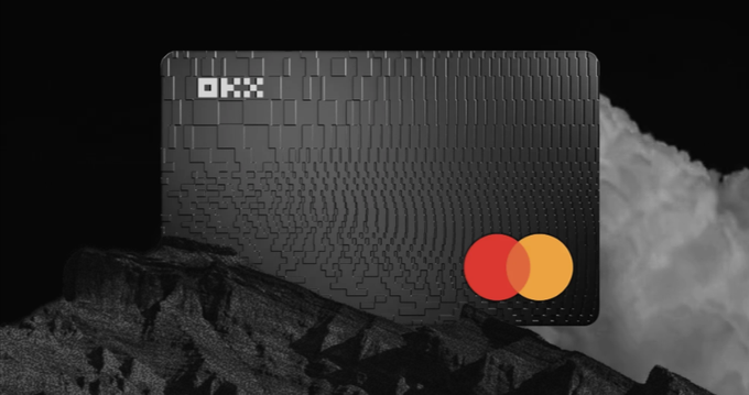 A black OKX credit card with a textured 3D surface, displayed on a dark rock with white smoke in the background.