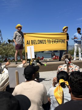 At an outdoor waterfront gathering, two people wearing yellow hats hold a large yellow banner reading "AI BELONGS TO EVERYONE," addressing a crowd of onlookers in a rally-like setting, evoking the inclusive spirit behind the "fomo" sentiment of not wanting to miss out on AI's future.