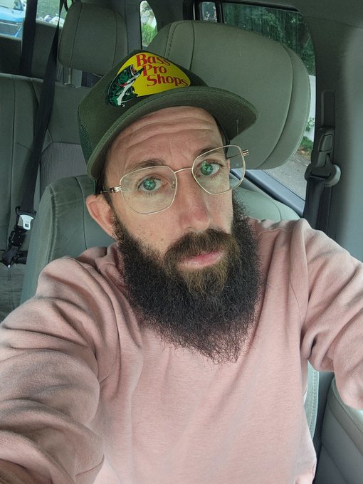 A man with a full dark beard, wearing gold-rimmed glasses and a green Bass Pro Shops trucker hat, takes a selfie inside a vehicle while wearing a pink sweater.