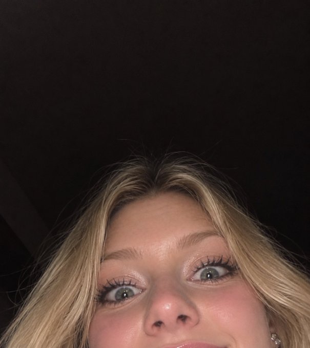 A close-up selfie captures a blonde woman's eyes, nose, and lips against a dark background, showcasing her makeup with shimmery eyeshadow and glossy lips.