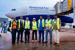 The image shows a group of people standing next to an airplane at an airport. There are six individuals, with four men wearing yellow vests and two women dressed in business attire. The airplane has the name "Rwandair" written on its side. In the background, there is another airplane visible as well.