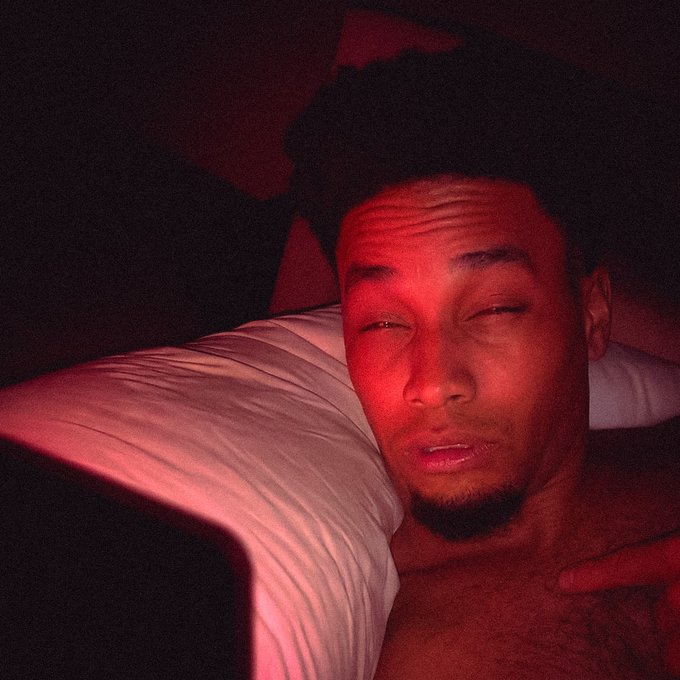 A man lies in bed with a tired, half-awake expression, lit by a dim reddish light, matching the groggy, just-woke-up vibe of a "gm wtf" morning post referencing $ANSEM.
