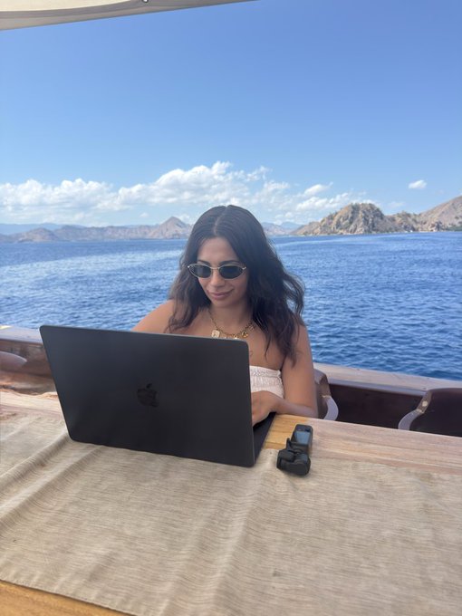 A woman in sunglasses sits at a waterfront table on a boat, working on a MacBook with a small action camera beside her, with scenic islands and blue ocean in the background—capturing the "remote work meets tropical escape" lifestyle the post evokes.