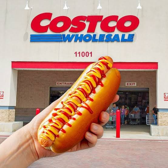 A hand holds up a massive hot dog with ketchup in front of a Costco Wholesale store entrance, with the store number '11001' visible above the doors.